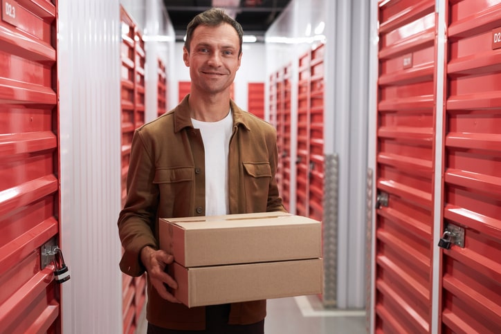  a man standing in a hallway of a storage facility with bright red roll-up doors. He is smiling and holding two cardboard boxes. The setting suggests he is either storing or retrieving items from one of the storage units.