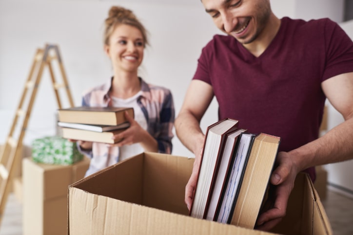 two people packing books into a cardboard box. The man in the foreground is placing books into the box, while the woman in the background is holding more books, smiling. They appear to be in the process of moving or organizing, with boxes and a ladder visible in the background.