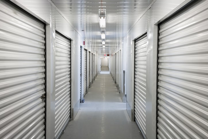 a long hallway in a storage facility, with rows of identical metal roll-up doors on either side. The corridor is well-lit by fluorescent ceiling lights, and the walls and floors are clean and light-colored.